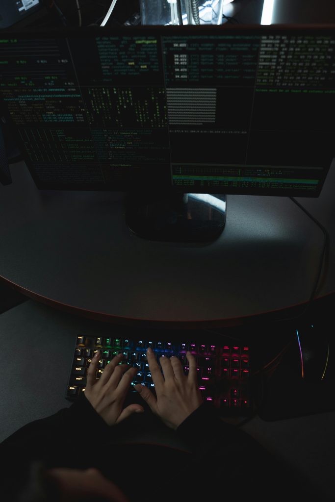 Hands typing on a rainbow-lit keyboard with code displayed on dual monitors.